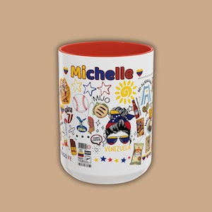 May include: A white ceramic mug with a red interior, featuring the name "Michelle" in red script. The mug is decorated with colorful Venezuelan-themed illustrations, including a baseball, sun, and the word "Venezuela".