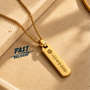 May include: A gold-toned metal pendant necklace featuring a rectangular bar charm with an engraved Spotify code and logo. The pendant hangs from a matching gold-toned box chain. The design is sleek, polished, and metallic in appearance.