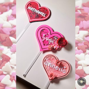 May include: Three heart-shaped cake toppers with clear stems. The top heart is red with the name "NATHANIEL" in white. The middle heart is pink with the name "Isabella" and a red paper rose. The bottom heart is pink with the name "annhella".