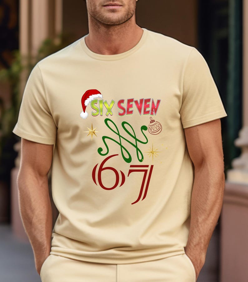 May include: A beige t-shirt with a festive design. The shirt features the words "SIX SEVEN" in red, green, and yellow, with a Santa hat on the "SIX". Below are the numbers "6 7" in red, with green swirls and gold stars.