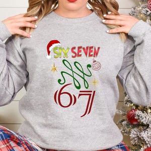 May include: A light gray sweatshirt with a festive Christmas design. The design includes the words "SIX SEVEN" in red and green, a Santa hat, a Christmas ornament, and the numbers "6 7".