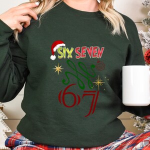 May include: Dark green sweatshirt with the text "SIX SEVEN" in red and green, with a Santa hat above the word "SIX". The design includes a Christmas ornament and starbursts. A white mug is held in the hand.