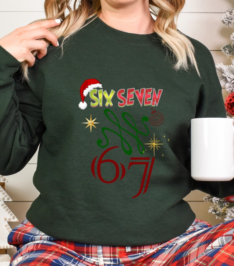 May include: Dark green sweatshirt with the text "SIX SEVEN" in red and green, with a Santa hat above the word "SIX". The design includes a Christmas ornament and starbursts. A white mug is held in the hand.