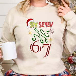 May include: A cream-colored sweatshirt with a festive design. The design includes the words "SIX SEVEN" in red and green, a Santa hat, and a Christmas ornament. The number "67" is also displayed in red.
