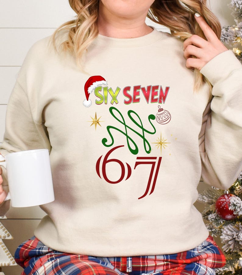 May include: A cream-colored sweatshirt with a festive design. The design includes the words "SIX SEVEN" in red and green, a Santa hat, and a Christmas ornament. The number "67" is also displayed in red.
