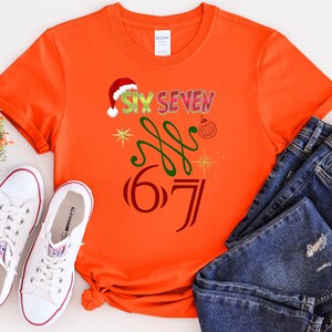May include: Orange t-shirt with the text "SIX SEVEN" in a festive design, including a Santa hat, green swirls, and a Christmas ornament. The number "67" is also displayed. White sneakers and blue jeans are also in the image.