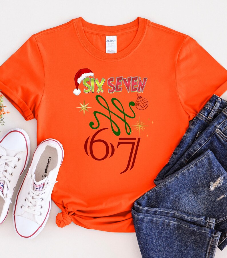 May include: Orange t-shirt with the text "SIX SEVEN" in a festive design, including a Santa hat, green swirls, and a Christmas ornament. The number "67" is also displayed. White sneakers and blue jeans are also in the image.