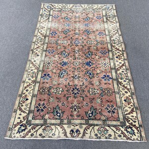 May include: Rectangular rug with a faded, vintage look. The rug features a central field of muted rose with floral and leaf motifs in blue, green, and white. A cream-colored border with intricate patterns frames the central design. The rug is likely made of wool.