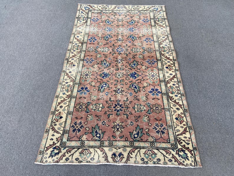 May include: Rectangular rug with a faded, vintage look. The rug features a central field of muted rose with floral and leaf motifs in blue, green, and white. A cream-colored border with intricate patterns frames the central design. The rug is likely made of wool.