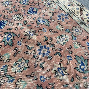 May include: Close-up of a vintage rug with a distressed, faded appearance. The rug features a floral pattern in shades of pink, blue, and cream. The design includes stylized flowers and leaves, with a border along the edge.