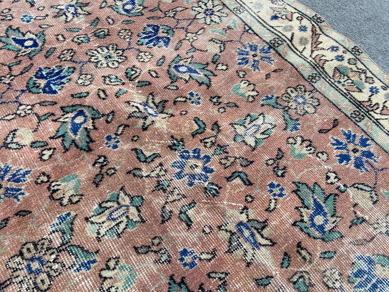 May include: Close-up of a vintage rug with a distressed, faded appearance. The rug features a floral pattern in shades of pink, blue, and cream. The design includes stylized flowers and leaves, with a border along the edge.