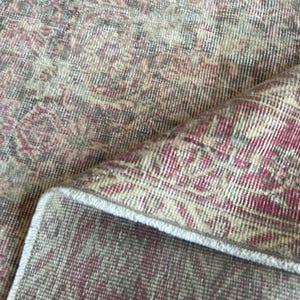 May include: A woven area rug featuring a faded, intricate floral and geometric pattern in shades of deep red, muted gray, and cream. The rug is folded over to reveal the textured pile on the top surface and the woven backing on the underside.