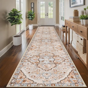 Puede incluir: Una alfombra rectangular larga con un diseño ornamentado y tradicional. La alfombra presenta un fondo color crema con un patrón repetido de formas florales y geométricas en tonos marrones, azules y naranjas. La alfombra está colocada en un pasillo con piso de madera.