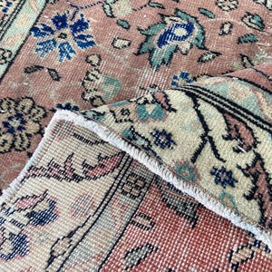 May include: Close-up of a vintage rug with a distressed, faded appearance. The rug features a floral pattern in shades of pink, cream, blue, and teal. The edges are frayed, adding to its antique charm. The rug is folded over, revealing the underside.