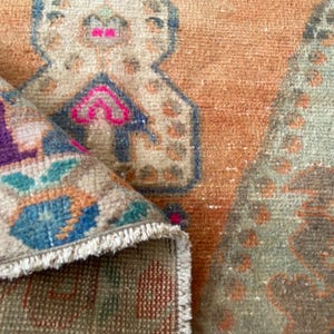 May include: A woven rug featuring a folded corner that reveals a vibrant underside with purple, blue, and pink geometric patterns. The main surface displays a warm orange background with stylized, faded motifs in muted green, brown, and cream tones.