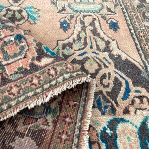 May include: Close-up of a rolled-up, vintage-style rug with a detailed, floral pattern. The rug features a color palette of muted peach, brown, teal, and green. The edges have a fringed border.