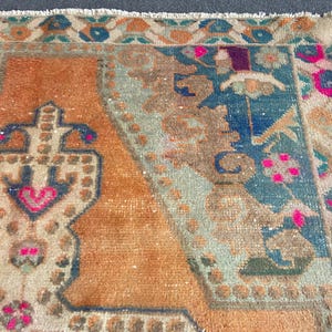 May include: A rectangular woven rug featuring a central orange field with a stylized geometric motif in dark blue and bright pink. The border displays a repeating pattern of floral shapes in shades of blue, orange, and pink with a fringed edge.