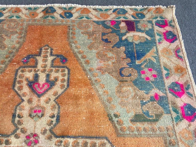 May include: A rectangular woven rug featuring a central orange field with a stylized geometric motif in dark blue and bright pink. The border displays a repeating pattern of floral shapes in shades of blue, orange, and pink with a fringed edge.