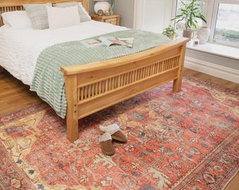 May include: A large rectangular area rug featuring a distressed pattern in shades of coral, red, cream, and muted blue. The rug is positioned on a wooden floor beneath a wooden bed frame with a white comforter and a light green textured throw blanket.