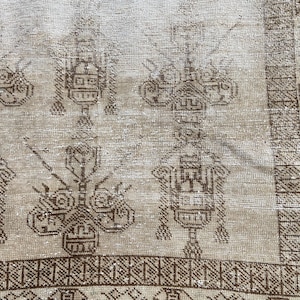 May include: A woven textile featuring a light beige background with dark brown geometric patterns. The design includes repeating symmetrical motifs and a detailed border with linear and circular shapes, creating a textured surface appearance.