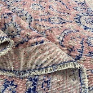 May include: A rolled-up, vintage-style rug with a distressed appearance. The rug features a faded pink background with intricate blue and cream-colored floral patterns. The edges have a fringed border, adding to its antique aesthetic.