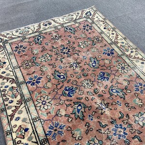 May include: A vintage rug with a distressed appearance. The rug features a central field of faded rose color with a floral pattern in blue, green, and white. The border is cream-colored with a repeating geometric design.