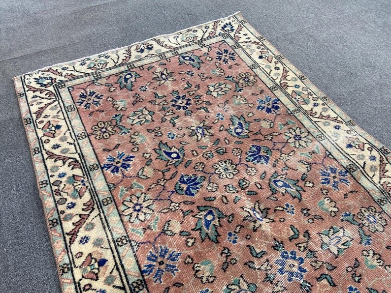 May include: A vintage rug with a distressed appearance. The rug features a central field of faded rose color with a floral pattern in blue, green, and white. The border is cream-colored with a repeating geometric design.
