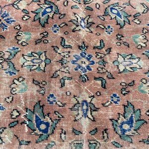 May include: Close-up of a vintage rug with a distressed, faded appearance. The rug features a repeating floral pattern in shades of pink, blue, and teal. The design includes stylized flowers and leaves, creating a classic, aged aesthetic.