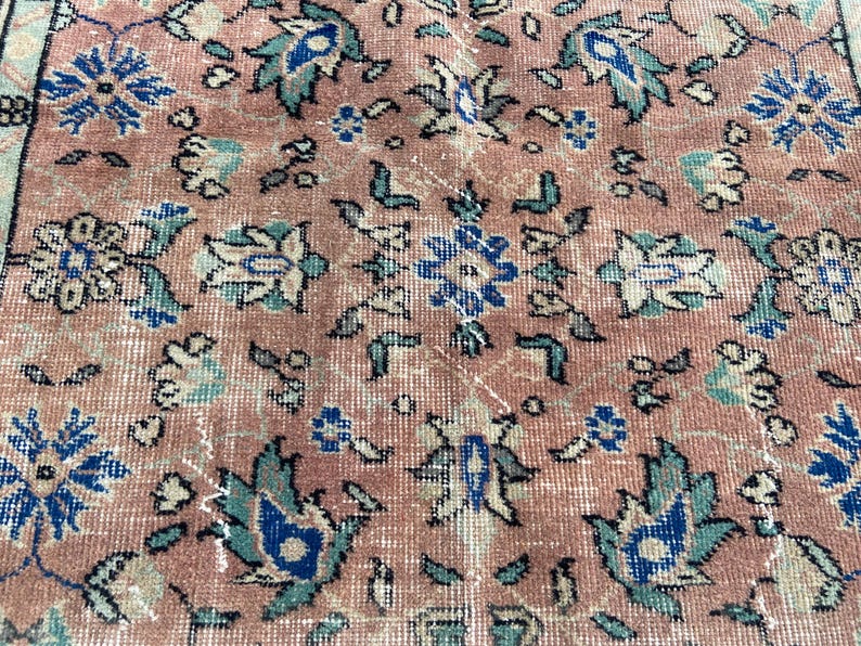 May include: Close-up of a vintage rug with a distressed, faded appearance. The rug features a repeating floral pattern in shades of pink, blue, and teal. The design includes stylized flowers and leaves, creating a classic, aged aesthetic.