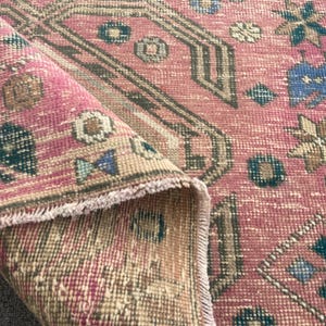 Könnte beinhalten: Vintage Teppich mit einem verblassten, abgenutzten Aussehen. Der Teppich hat eine rosa-beige Farbpalette mit geometrischen und floralen Mustern in Türkis, Braun und Creme. Die Ränder sind ausgefranst, was seinen antiken Charme unterstreicht.