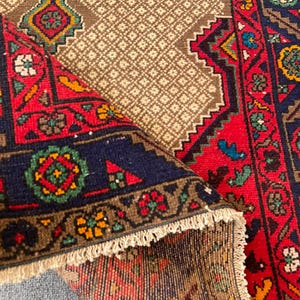May include: A woven rug featuring a folded corner that reveals a tan center with a repeating diamond grid pattern. The border displays intricate geometric motifs in vibrant red, deep blue, green, and yellow, finished with a fringe edge.