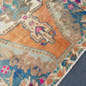 May include: A rectangular woven rug featuring a central orange field with a geometric medallion pattern in shades of blue, beige, and brown. The design includes intricate floral motifs and small accents of bright pink throughout the textile.