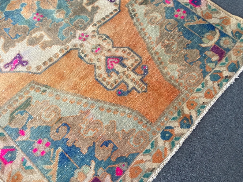 May include: A rectangular woven rug featuring a central orange field with a geometric medallion pattern in shades of blue, beige, and brown. The design includes intricate floral motifs and small accents of bright pink throughout the textile.