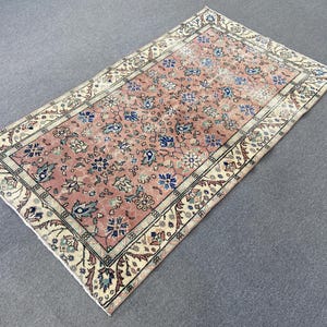 May include: A rectangular, vintage-style rug with a faded, floral pattern. The rug features a central field of muted pink with blue and white floral motifs. A cream-colored border with intricate designs frames the rug, creating a classic aesthetic.