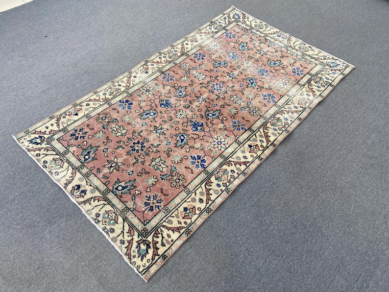 May include: A rectangular, vintage-style rug with a faded, floral pattern. The rug features a central field of muted pink with blue and white floral motifs. A cream-colored border with intricate designs frames the rug, creating a classic aesthetic.