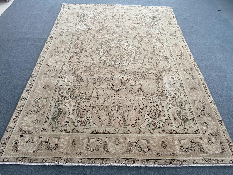 May include: A rectangular, beige-toned rug with an intricate, floral-inspired central medallion design. The rug features a detailed border with repeating floral patterns and a faded, vintage aesthetic. The rug is placed on a blue surface.