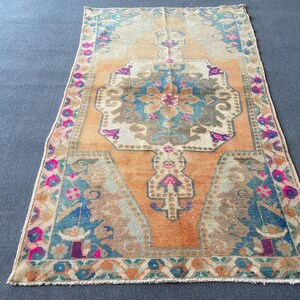 May include: A rectangular area rug featuring a central medallion in shades of blue, beige, and magenta set against a warm orange background. The border displays a repeating floral pattern in pink, blue, and green, with fringed edges on both ends.
