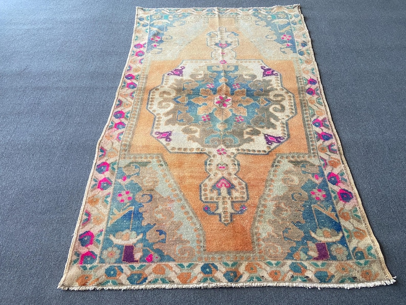 May include: A rectangular area rug featuring a central medallion in shades of blue, beige, and magenta set against a warm orange background. The border displays a repeating floral pattern in pink, blue, and green, with fringed edges on both ends.