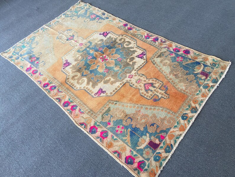 May include: A rectangular area rug featuring a central medallion in shades of blue and beige set against a vibrant orange background. The border displays intricate floral patterns in pink, teal, and brown, finished with short white fringe at ends.