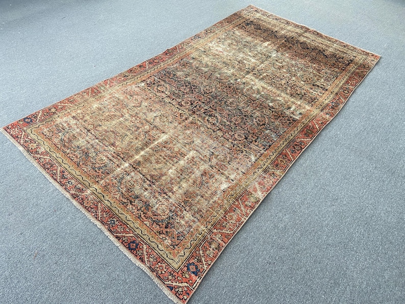 May include: A rectangular, distressed rug with a central field of brown and beige tones. The rug features a detailed, faded floral pattern and a border in shades of red, navy, and gold. The rug's design suggests a vintage or antique style.