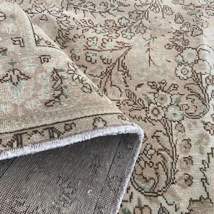 May include: Beige and brown vintage rug with floral and geometric patterns. The rug is partially rolled up, revealing a fringed edge and a detailed border. The design features muted tones and a distressed appearance, suggesting an antique or traditional style.