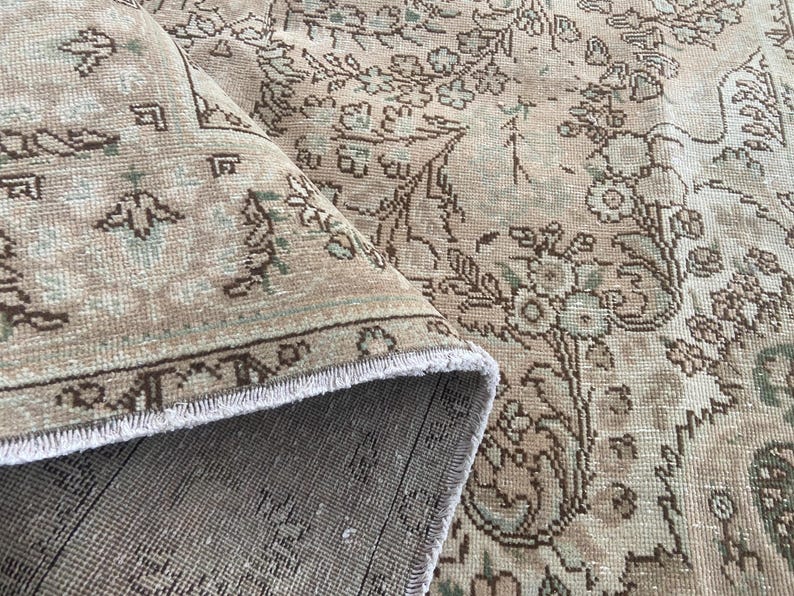 May include: Beige and brown vintage rug with floral and geometric patterns. The rug is partially rolled up, revealing a fringed edge and a detailed border. The design features muted tones and a distressed appearance, suggesting an antique or traditional style.