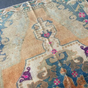 May include: A woven rug featuring a central tan field with a geometric medallion in shades of blue, brown, and cream. The border displays intricate patterns in teal, tan, and blue, accented with small, vibrant pink floral motifs throughout.