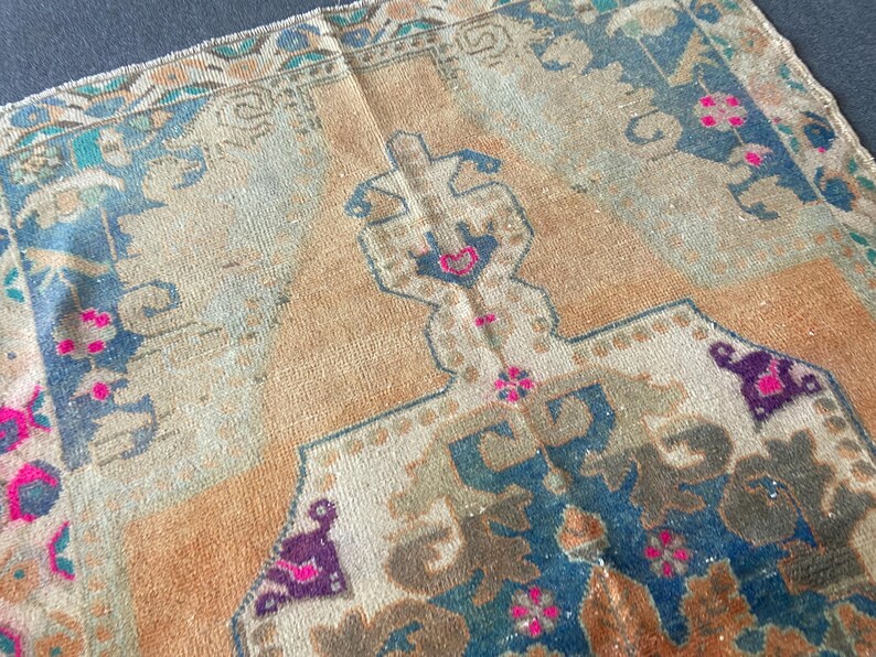May include: A woven rug featuring a central tan field with a geometric medallion in shades of blue, brown, and cream. The border displays intricate patterns in teal, tan, and blue, accented with small, vibrant pink floral motifs throughout.