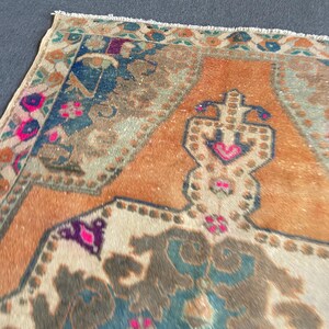 May include: A rectangular area rug featuring a central orange field with a large, intricate geometric medallion in shades of blue, brown, and cream. The border displays floral motifs in vibrant pink, deep blue, and green with a fringed edge.