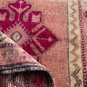 May include: A woven rug featuring a vibrant fuchsia and light pink geometric pattern with a central medallion. The rug is folded over to reveal a dark, textured underside with visible fibers and a dense, knotted pile construction throughout.