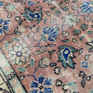 May include: Close-up of a vintage rug with a faded, distressed appearance. The rug features a repeating floral pattern in shades of dusty rose, blue, and cream. The design includes stylized flowers and leaves, with a border along the edge.