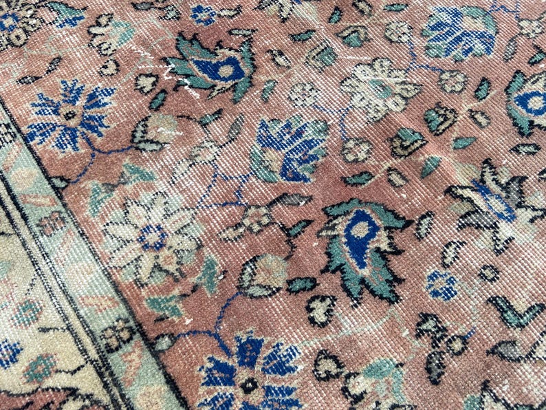 May include: Close-up of a vintage rug with a faded, distressed appearance. The rug features a repeating floral pattern in shades of dusty rose, blue, and cream. The design includes stylized flowers and leaves, with a border along the edge.