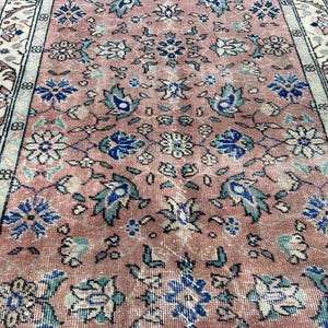 May include: A vintage rug with a distressed, faded appearance. The rug features a central field of muted rose with floral and foliate patterns in blue, teal, and cream. The border is cream with floral motifs.