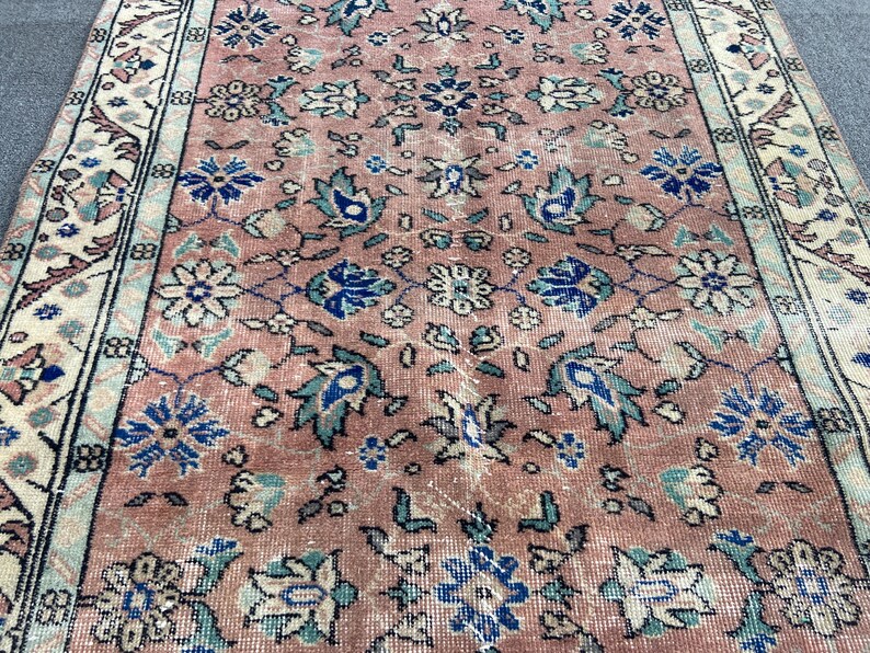 May include: A vintage rug with a distressed, faded appearance. The rug features a central field of muted rose with floral and foliate patterns in blue, teal, and cream. The border is cream with floral motifs.